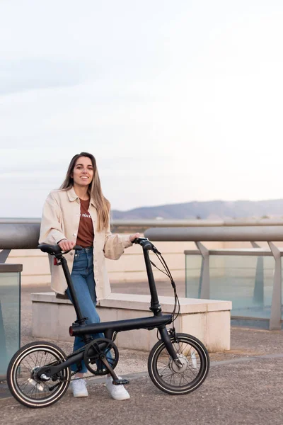 Vue verticale de la femme heureuse et souriante à la caméra tenant un vélo électrique dans la ville avec espace de copie — Photo