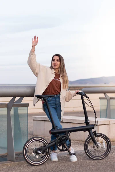 Vertical view of young woman waving at someone whle holding an electric bicycle for going back home from the city center — Photo
