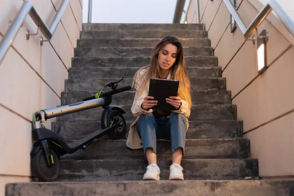 Frau sitzt auf Stufen und benutzt ein Tablet, auf ihrer Seite liegt ein Elektroroller. Mit Nachtlicht und Scheinwerfern — Stockfoto