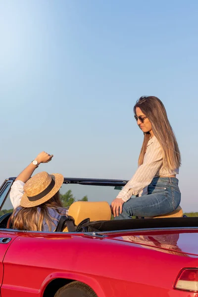 Dwie młode kobiety cieszące się czasem spędzonym razem na czerwonym klasycznym, kabrioletowym samochodzie podczas wakacji — Zdjęcie stockowe