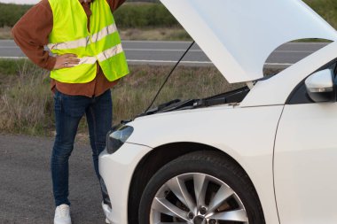 Yolun kenarında yelek giymiş tanınmayan bir adam kaputu açık bir şekilde arabasında kırılanı bulmaya çalışıyor.