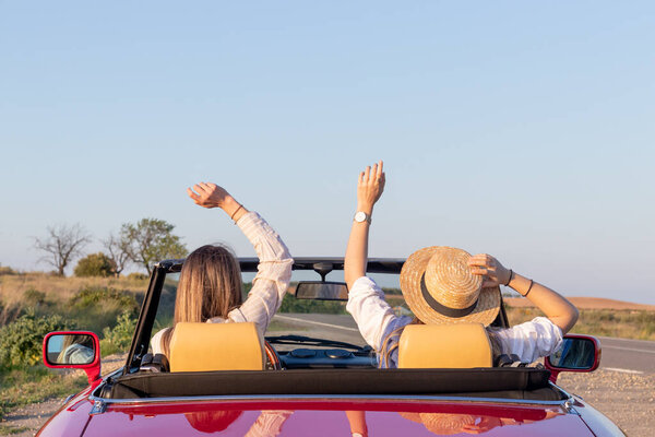 Happy free girlfriends with straw hat driving in red retro car cheering joyful with arms raised. Road trip travel concept. With copy space under blue sky