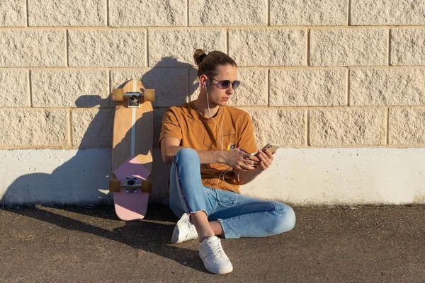 Young hipster man on a bun hairstyle sitting on the floor with a skate board on the side listening to music on the smartphone