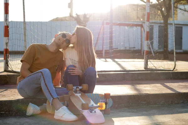 Cute lovely heterosexual couple sitting kissing on forehead with sun behind them after going on long board — Fotografia de Stock