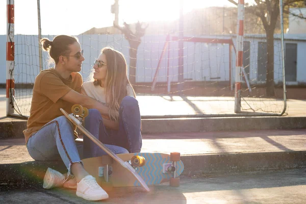In love couple looking to each other sitting in a park with two skate boards and sunset sun behind them — Fotografia de Stock