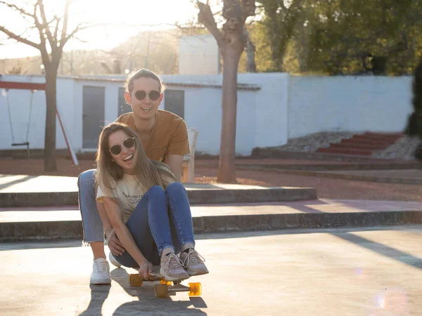 Summer love between a couple of teenagers having a good time on a skate board in the city park at sunset — Fotografia de Stock