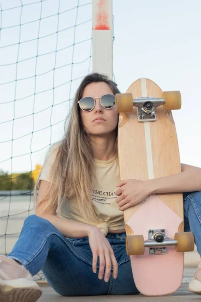 Přední záběr vzdorné blondýny dívka sedí na podlaze vedle její růžové vintage skateboard — Stock fotografie