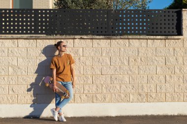 Beyaz bir duvara yaslanmış, elinde kaykay kaykayıyla bahar güneşinin tadını çıkaran rahat bir adam.