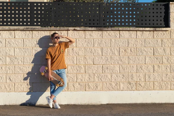 Kaykaycı adam gün batımından önce güneş ışığına bakan beyaz bir duvara karşı uzun kaykayı tutarak rahatladı.