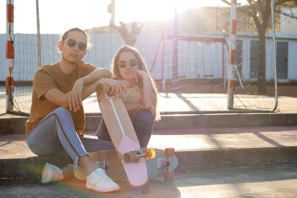 Two hipster friends sitting on steps leaning on cute skate board in a spring afeternoon with sunlight behind them — Fotografia de Stock