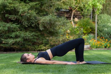 Forma girmek için yazın bahçede egzersiz yapan genç bir kadının yan görüntüsü. Çimenlerdeki siyah bir paspasın üzerinde