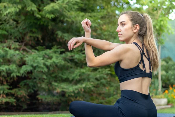 Vista lateral de una joven atractiva sentada en el jardín calentándose para la rutina de ejercicios de yoga — Foto de Stock