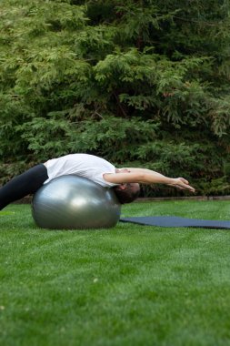 Dikey olarak, yoga yapan genç bir adamın rahatlamak için bahçedeki denge topunda esneme hareketleri yaptığını görüyorum.
