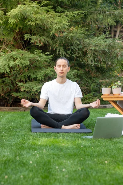 Vista frontal del joven con moño practicando yoga de loto posan en el jardín con una clase de curso en línea de ordenador — Foto de Stock