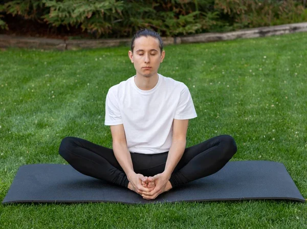 Joven en posición de loto mientras practica yoga con los ojos cerrados sobre una alfombra negra en el jardín — Foto de Stock