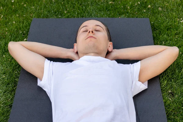 Hombre joven en camisa blanca con los ojos cerrados y los brazos abiertos descansando sobre una esterilla de yoga mirando al cielo en el jardín — Foto de Stock