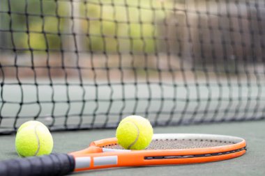 İzole edilmiş turuncu tenis raketi ve toplar arka planda ağ ile sahada dinleniyor.