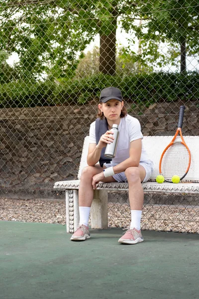 Mladý muž odpočívající na lavičce po tenisovém zápase s lahví vody v ruce a raketou — Stock fotografie
