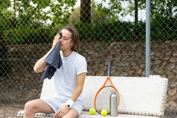 Egy fiatalember törölközővel törölgeti az izzadságát egy padon ülve, miután teniszezett a pályán. — Stock Fotó