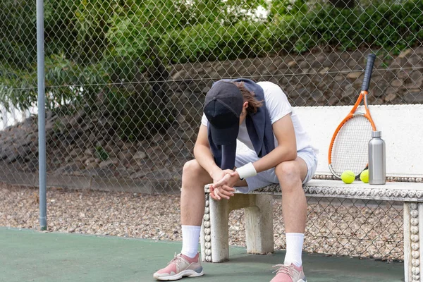 Exhausted sportsman sitting on a bench after tennis training class looking down to the floor in the court