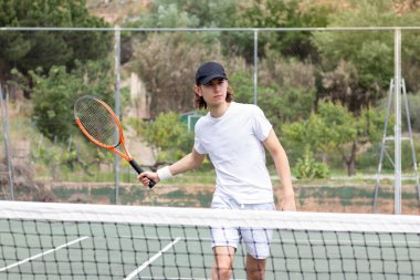 Uzun saçlı, şapkalı genç bir adam kortta ortasında ağ olan bir tenis maçında oynuyor. Topa vurmaya çalışıyorum.