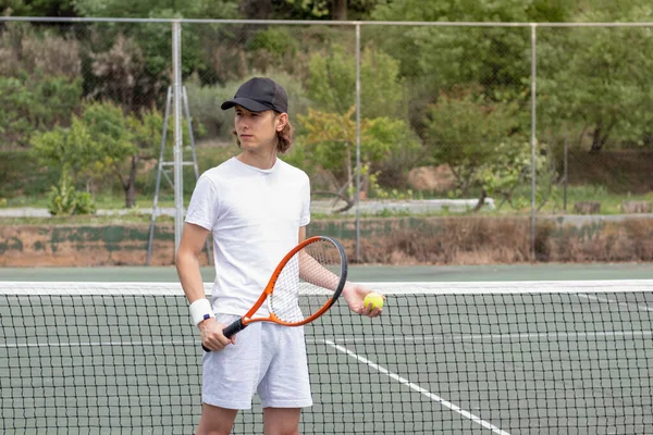 Şapkalı, elinde tenis ekipmanı olan ve yeşil kortta oynamayı bekleyen genç bir çocuğun portresi.. — Stok fotoğraf