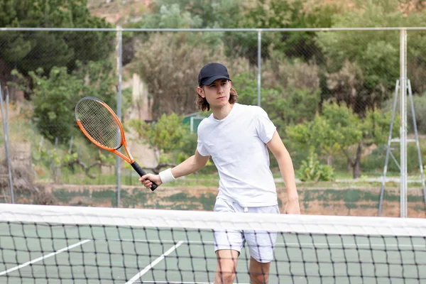 Uzun saçlı, şapkalı genç bir adam kortta ortasında ağ olan bir tenis maçında oynuyor. Topa vurmaya çalışıyorum. — Stok fotoğraf