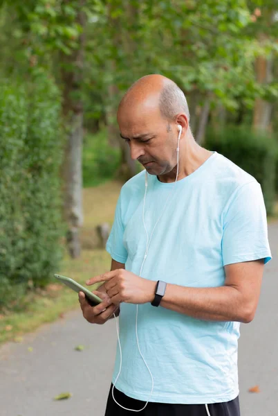 Orta boy yetişkin bir adam cep telefonuyla müzik dinlerken doğa arasında spor yaparken çekilmiş.