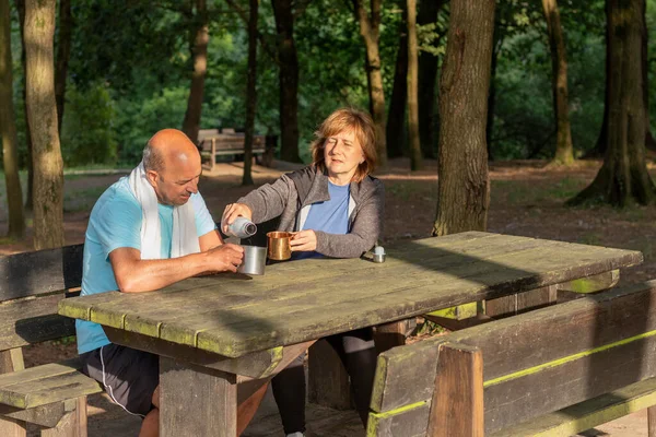 Senior couple sitting in a picnic table in the forest drkinking water from bottles in metal camping cups in the afternoon