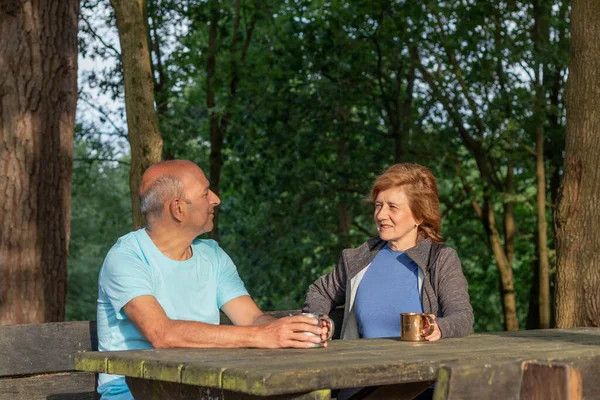 Senior couple looking to each other and smiling while sitting on a picnic wooden table resting in the woods