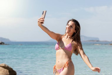Akdeniz 'de renkli bir bikini mayosunun arka planında kumsalda selfie çeken güzel, genç esmer kız.