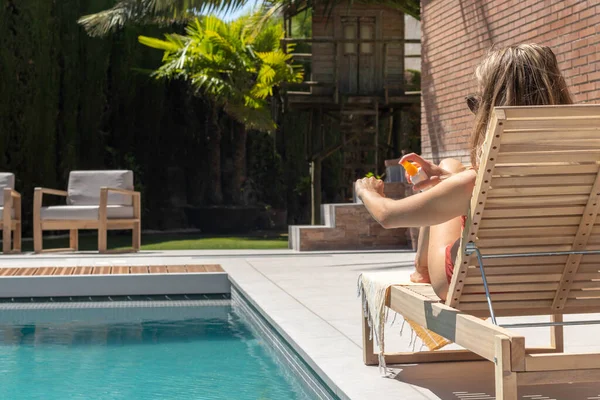 Kaydedilemez genç sarışın kadın bahçedeki havuzda güneşlenirken koluna güneş kremi sürüyor.