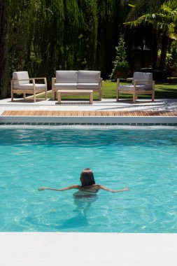 Vertical view of backward unrecognizable young woman in the middle of the swimming pool with lounge area in the background in summer