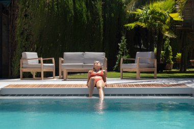 Front view of young woman laying down on the edge of the swimming pool area with wooden loungers in the back garden