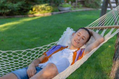 Young man with eyes closed sitting in a hammock listening to music with a sun light in face during summer day