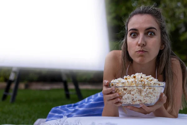 Portrait of a young woman with her eyes open and a face of surprise. as her mouth is full of popcorn at the summer cinema in the park