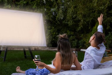 Happy unrecognizable couple sitting in the grass watching a movie in the garden holding red wine glasses