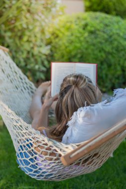 Yazın hamakta otururken arkada kitap okuyan tanınmayan genç bir kadının dikey görüntüsü.