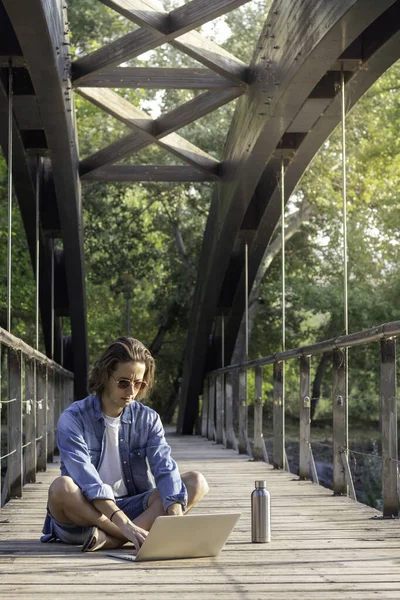 Baharda gün batımında ahşap bir köprünün zemininde oturan genç çevrimiçi işçinin dikey görüntüsü doğayla iç içe.