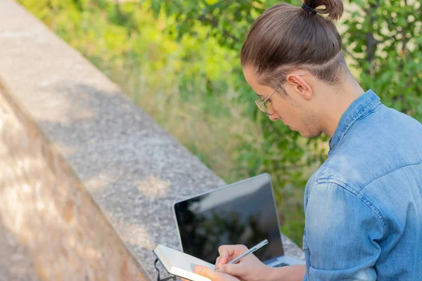 Back view of young man on a bun taking notes to create a new business online using his laptop sitting in a park in europe