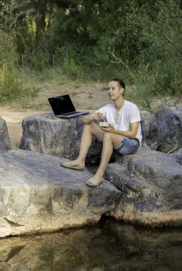 Dijital göçebe genç erkek, yeşil bir nehrin yanında piknik yapmak için mola veriyor.