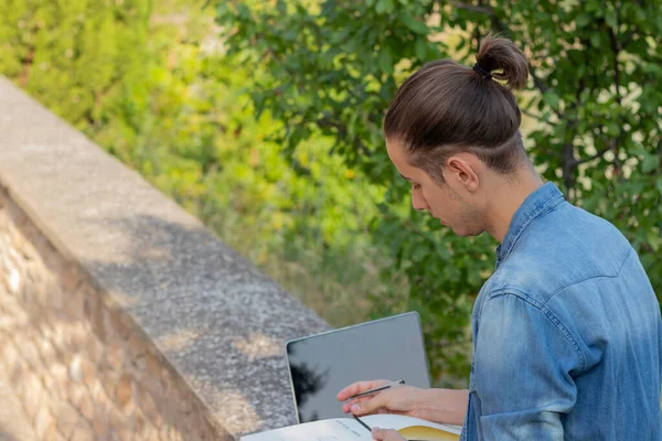 Young man on bun hairstyle looking down to his notebook enjoying leisure activity in the nature with laptop computer