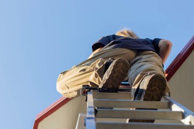 Bottom view of unrecognizable young working woman stepping on a ladder going into the roof with blue clear sky background
