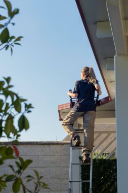 Vertical back view of unrecognizable young woman on ponytail climbing a ladder with plants around while working on her daily job
