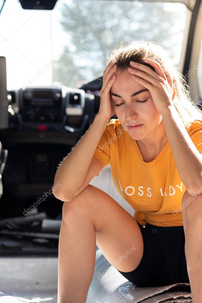 Young woman with headache and hands on head while converting her DIY camper sitting on the isolation roll with backlight in summer