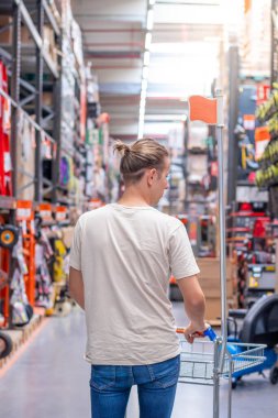 Endüstriyel hırdavat dükkanının koridorunda alışveriş arabasını iten genç adamın dikey görüntüsü bulanık arka planı