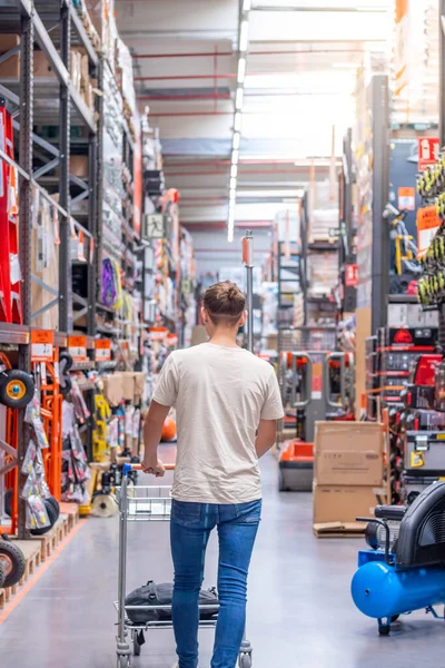 Young man on a bun buying and walking throw the middle of the aisle of a hardware store with a trolley in hands