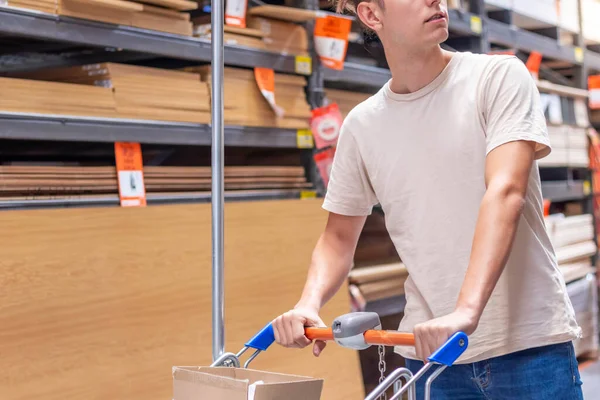 Unrecognizable young man carrying a shopping trolley in a hardware store with wood stored in the blurred background