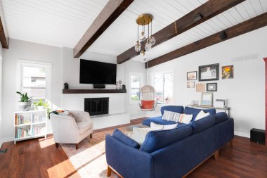 ELMHURST, IL, USA - SEPTEMBER 2, 2020: A renovated modern farmhouse living room with a blue couch, white fireplace, wood beams on a white shiplap ceiling, and a television mounted above the fireplace. clipart