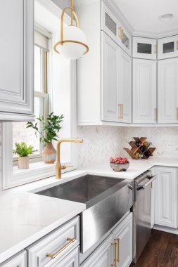 ELMHURST, IL, ABD - 2 Temmuz 2020: Altın kolye ışığı, paslanmaz çelik önlük lavabo, beyaz dolaplar ve herringbone mermer arka sıçrama ile çekilen bir mutfak lavabosu ayrıntıları.